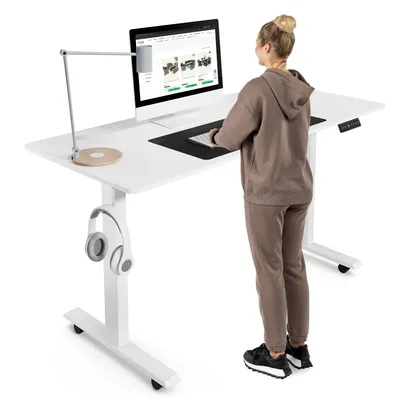 A woman is standing at a white adjustable desk, working on a computer. The desk has a lamp and headphones attached.