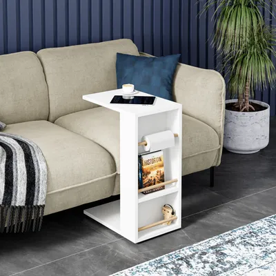 A white side table with shelves is placed next to a sofa in a living room setting. A roll of paper, a book, and a small figurine are on the shelves. A tablet and a cup of coffee are on the top surface of the table.