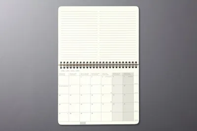 An open desk calendar with a monthly planner and lined note pages, laid flat on a gray surface.
