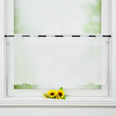 A white sheer curtain with embroidered leaf patterns hangs on a window rod, with two artificial sunflowers placed on the windowsill below.