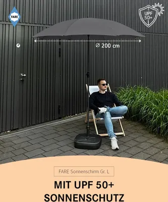 A man sits in a chair under a large gray umbrella outdoors, with a dark wall and greenery in the background.
