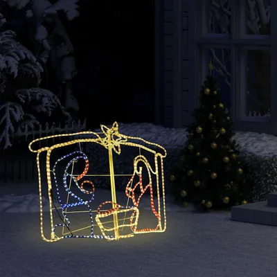 A lighted nativity scene decoration is displayed outdoors at night, with a Christmas tree in the background.