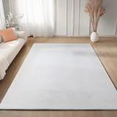 A white rug is laid out on a wooden floor in a living room setting, with a sofa and decorative vase visible.