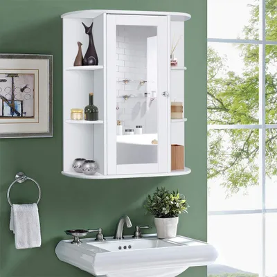 A white bathroom cabinet with a mirrored door is mounted on a green wall above a white sink. The cabinet has shelves with decorative items and toiletries. A towel hangs on a ring to the left of the sink, and a potted plant sits on the counter to the right.