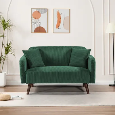 A green corduroy sofa with two throw pillows is shown in a well-lit room with abstract art on the wall and a floor lamp.