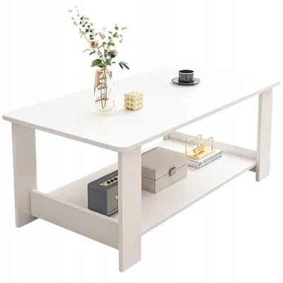 A white coffee table with a two-tier shelf, decorated with a vase of flowers, a decorative cube, a cup and saucer, a grey box, a small speaker, and books.