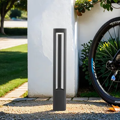 A modern gray aluminum LED bollard light stands on a gravel path next to a white wall and a bicycle wheel.