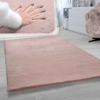 A close-up of a soft, pink, high-pile rug with a hand pressing into it, suggesting its plushness. The rug is shown on a light gray wooden floor.