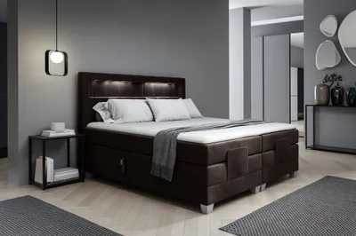 A bedroom scene featuring a large boxspring bed with integrated lighting, a bedside table, and decorative elements in the background.