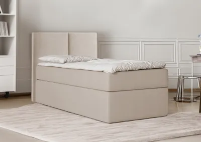 A beige upholstered bed with a headboard is shown in a room with a bookshelf and a chair in the background.