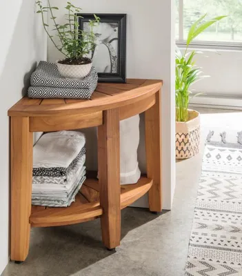 A wooden corner shelf unit with folded towels and a plant, placed in a room with a rug and a window.