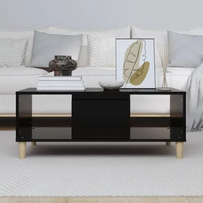 A modern black coffee table with wooden legs is placed in a living room setting, in front of a white sofa with decorative pillows. The table has a sleek, minimalist design with open shelving on the sides and a central drawer or cabinet. It is adorned with 