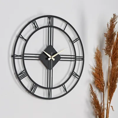 A black metal wall clock with Roman numerals is displayed on a white wall, next to some dried grass stalks.
