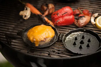 A burger patty with melted cheese is cooking in a cast iron pan on a grill, surrounded by grilled vegetables.