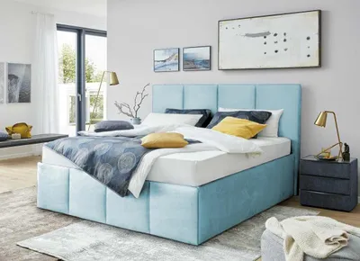 A bedroom scene featuring a blue upholstered bed with storage, bedside table, lamp, and artwork.