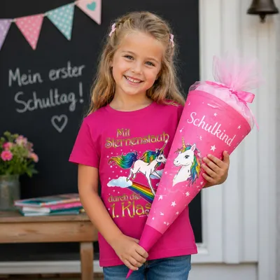Fröhliches Mädchen trägt pinkes Einhorn T-Shirt „Mit Sternenstaub durch die 1. Klasse“ und hält eine Zuckertüte zur Einschulung.