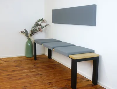 A modern bench with gray seat cushions and black legs, placed in a room with wooden flooring and a green vase with dried plants.