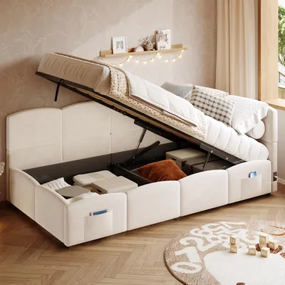 A beige upholstered bed with its storage compartment open, revealing stored items. The bed is in a room with a wooden floor and a window with curtains.