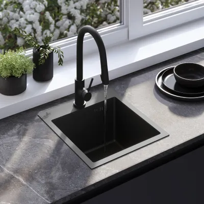 A black kitchen sink with a black faucet is shown with water running into it. The sink is on a gray countertop next to a window with white flowers outside. There are two small potted plants on the windowsill.
