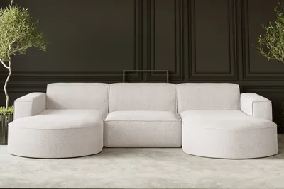 A modern U-shaped sofa in a light ecru fabric is shown in a minimalist room with dark paneled walls and a light gray floor. Two potted plants flank the sofa.