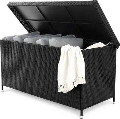 An open black wicker storage box filled with gray cushions and a white blanket draped over the side, set against a white background.