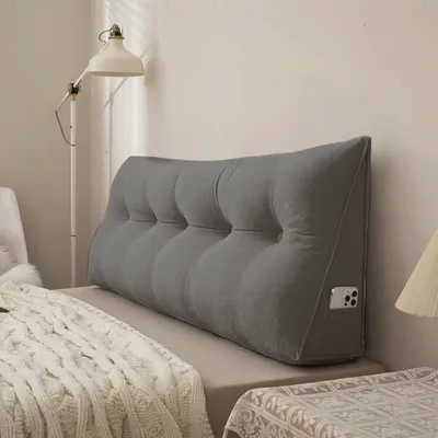A grey wedge pillow is placed on a bed against a wall, with a lamp and a blanket visible.