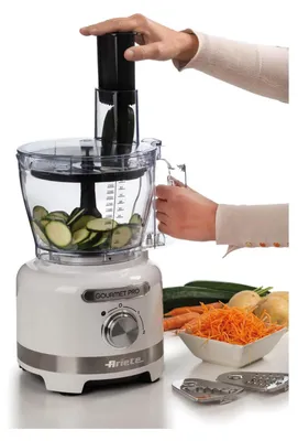 A white Ariete Gourmet Pro food processor is shown in use, with a person demonstrating how to insert a zucchini into the chute. Sliced zucchini fills the bowl, and shredded carrots, potatoes, and whole vegetables are visible nearby, along with food process