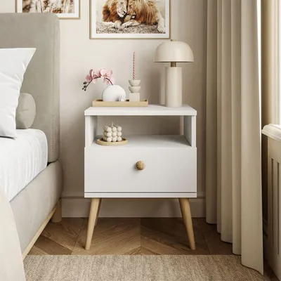A white nightstand with a drawer and an open shelf, decorated with a vase, a candle, and a lamp, placed next to a bed.