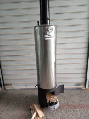 A stainless steel wood-fired water heater boiler with firewood in front of it, placed against a corrugated metal wall.