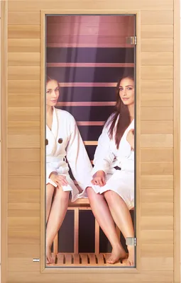 Two women in white robes sitting inside a wooden sauna with a glass door.