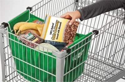 A hand is placing a box of cereal into a green shopping cart bag inside a shopping cart.