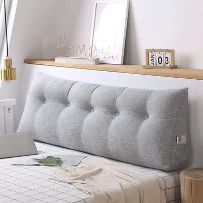 A bed with a large grey wedge pillow against the headboard, with decorative items on a shelf above.