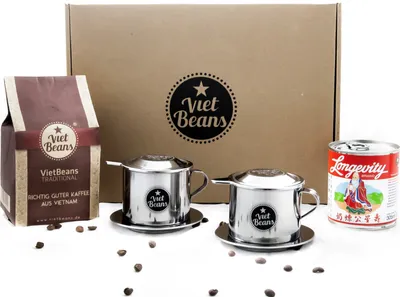 A coffee gift set including coffee beans, two Vietnamese coffee filters, and a can of condensed milk, arranged on a white background with a cardboard box in the background.