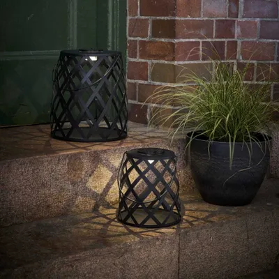 Two black metal lanterns with a diamond pattern are placed on stone steps next to a potted plant. The lanterns are lit, casting a patterned shadow on the steps. A green door is visible in the background.