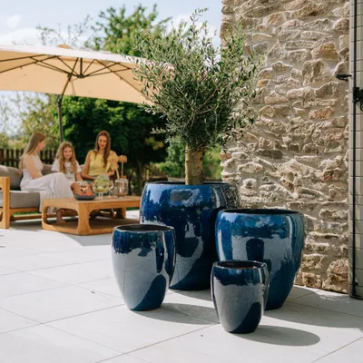 Three women are sitting on a sofa under a large umbrella, with several large blue planters in the foreground. A stone wall is in the background.