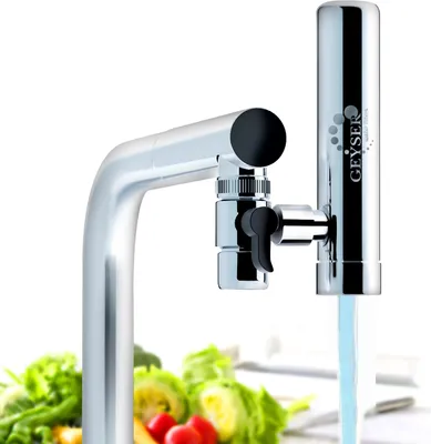 A water filter attached to a kitchen faucet, with water flowing through it and blurred vegetables in the foreground.
