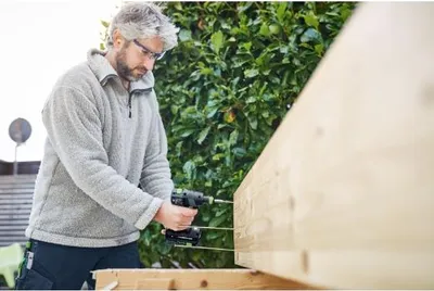 A man is using a cordless drill to screw into a wooden beam outdoors.