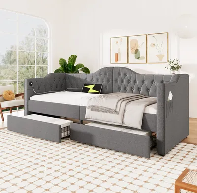 A grey upholstered daybed with two pull-out drawers, set up in a well-lit room with a large window and decorative artwork on the wall.