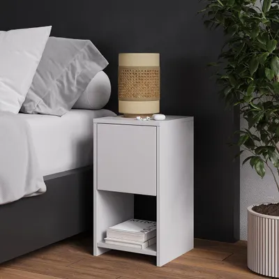 A bedside table with a lamp and books next to a bed, with a plant in the background.