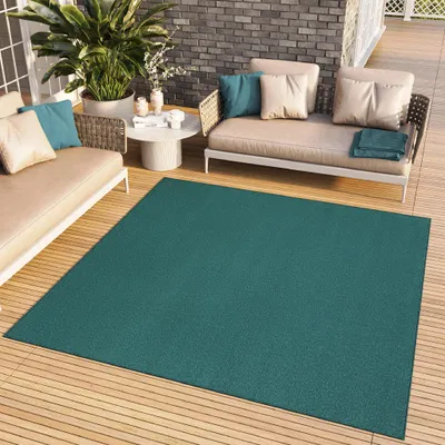 A teal rug is placed on a wooden deck next to outdoor furniture, including a sofa and chairs, with plants and a coffee table.