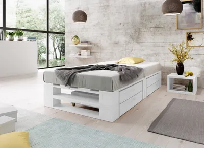 A bedroom scene with a white wooden bed frame with drawers, a mattress, and bedding. Large windows with a view of trees are in the background.