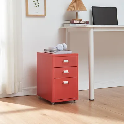 A red metal roll container with three drawers sits on a wooden floor next to a white desk. There are headphones and books on top of the container. A laptop and a lamp are on the desk.