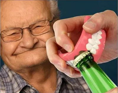 An elderly man with glasses is smiling as a hand holds a pair of dentures to open a green bottle.
