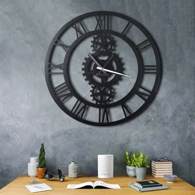 A large, black, gear-themed wall clock with Roman numerals hangs on a textured gray wall above a wooden desk. The desk has various decorative items and books.