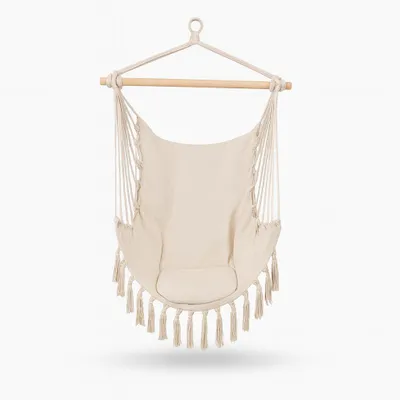 A cream-colored hammock chair with tassels hanging from a wooden bar, suspended by ropes.