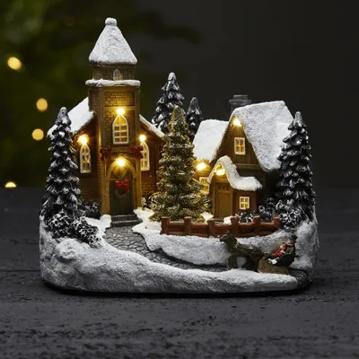 A miniature Christmas village scene with a church, houses, and trees, covered in snow and illuminated by warm LED lights.