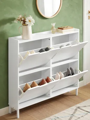 A white shoe cabinet with multiple flip-down compartments filled with shoes, placed against a green wall with a mirror and decorative items on top.