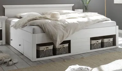 A white wooden double bed with storage drawers and baskets, featuring a white duvet and a straw hat on top.