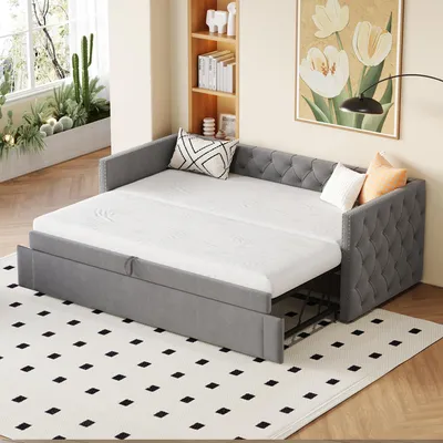A grey upholstered daybed with a pull-out trundle bed is set up in a living room, featuring a patterned rug and a painting on the wall.