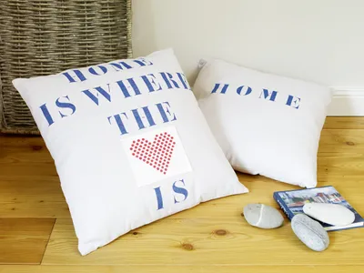 Two white throw pillows with blue lettering and a red heart design are placed on a wooden floor next to a book and some stones.
